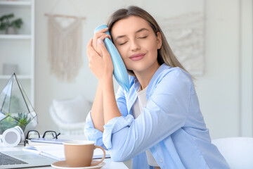 Young woman with laptop, cup of coffee and hot-water bottle feeling relief from tooth ache at home