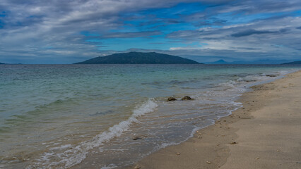 A secluded tropical beach. The waves of the turquoise ocean are foaming on the sand. Mountains in the distance against a background of blue sky and clouds. Madagascar.