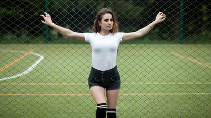 girl standing in front of a mesh fence on a sports field. She is wearing a white T-shirt and black high-waisted shorts.