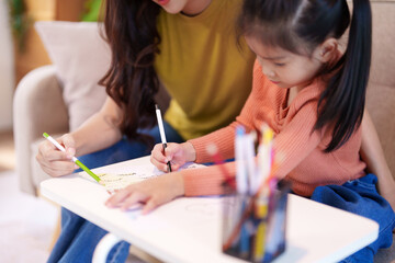 A mother assisting her daughter with homework at the table, surrounded by books and stationery,...