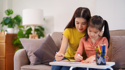 A mother assisting her daughter with homework at the table, surrounded by books and stationery,...