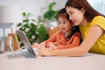A mother is teaching her young daughter to do homework together at the table, creating a supportive and educational environment.