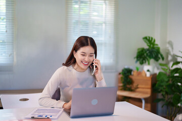 Obraz premium A woman is multitasking, talking on the phone while using her laptop and reviewing documents on the desk.