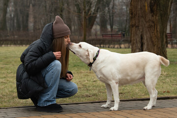 A woman in a dark coat and knitted hat kneels to share a sweet moment with her friendly white...