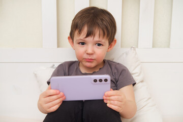 Naklejka premium A young child lying on a pillow and holding a phone in the bedroom. An adorable baby holding a smartphone while resting on a pillow in bed. Kid aged three (three-year-old )
