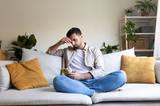 Worried and upset man looking at phone at home sitting on the sofa.