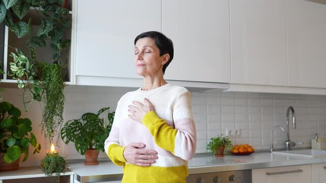 Relaxed mature woman practices breathing exercises at home, hands on chest and eyes closed, focused on deep breathing. Female mindfulness and calmness, tension release, stress relief at home alone
