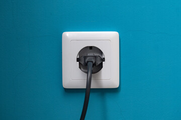Black power plug is plugged into a white socket on a cyan-painted wall.