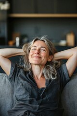 Obraz premium Photo of a middle-aged woman relaxing on a couch at home, happy and content with her arms behind her head, smiling and looking up in the distance. In a modern kitchen.