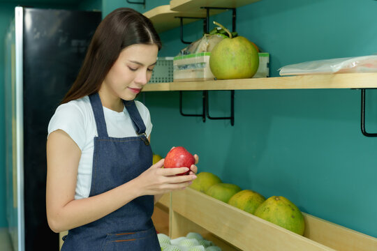 Young female shopkeeper inspecting fresh red apple with care in a modern fruit store. Focused on ensuring product quality and providing an attractive presentation for customers.