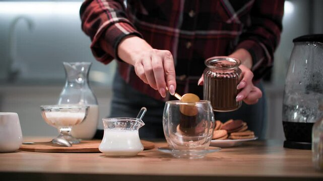 preparation of favorite drinks, female hand prepares cappuccino and pours coffee into transparent cup, pours boiling water and froths milk with cappuccino maker on kitchen table, close-up