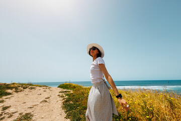 Summer mood. A young happy woman in a white dress and hat walks carefree along the seashore.