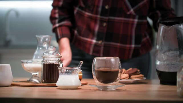 hot drink preparation, young woman preparing cappuccino and pouring boiling water over coffee in a glass cup and frothing milk with a cappuccino maker on the kitchen table, close-up