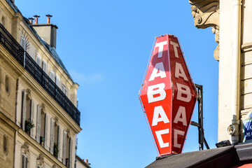 Red french tobacco store sign with white lettering and haussmannian building in the background against blue sky.