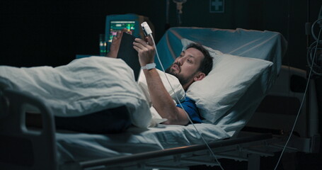 Sick male patient in hospital ward in bed drinks a glass pf water and holds tablet in hand checks...