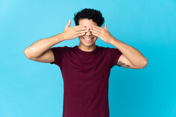 Young Venezuelan man isolated on blue background covering eyes by hands and smiling