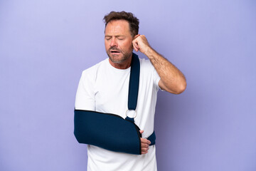 Middle age caucasian man with broken arm and wearing a sling isolated on purple background frustrated and covering ears