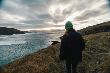 Chica rubia joven irreconocible caminando por la costa cerca del mar, gorro verde, prado con...