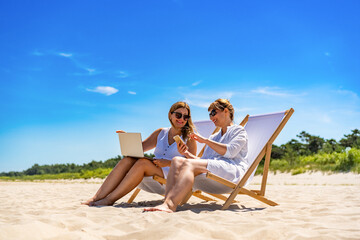 Remote work on beach. Two middle aged women sitting on deckchairs using laptop and smartphone on sandy beach on sunny day
