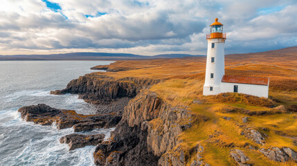 Naklejka premium high angle view of historic lighthouse standing on rugged coastal cliff, surrounded by dramatic ocean waves and golden grass under partly cloudy sky