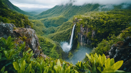 Fototapeta premium high angle view of majestic waterfall plunging into lush green valley surrounded by dense forest and misty mountains, evoking sense of tranquility and natural beauty