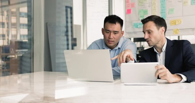 Two male coworkers using gadgets for working together at office table, comparing online data on tablet and laptop, discussing Internet marketing strategy, promotion on social media