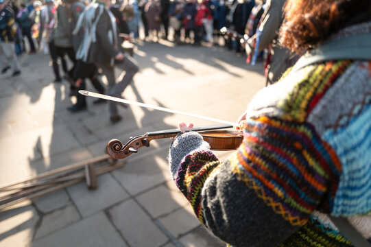 Female violinist seen with her instrument at a Morris Dancer golf festival in the UK. A band of adults can be seen dancing on a city street.