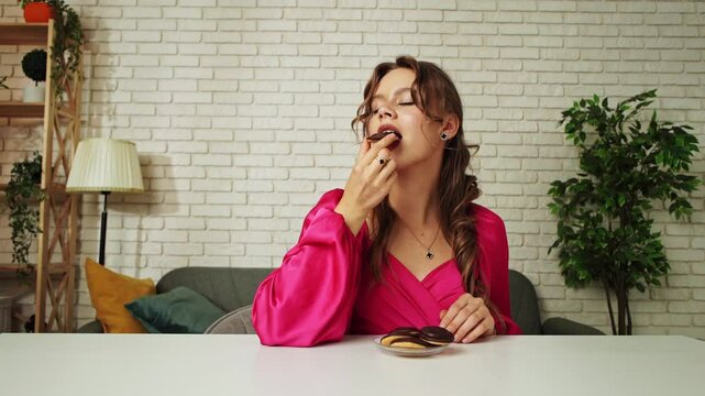 Young woman sitting at table and eating chocolate glazed cookie, taking a bite and chewing, enjoying the taste of sweet dessert snack, happy expression.