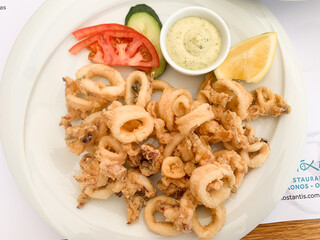 Top view of fried calamari rings served on a white plate with sliced tomato, cucumber, lemon wedge, and a small cup of creamy dip.