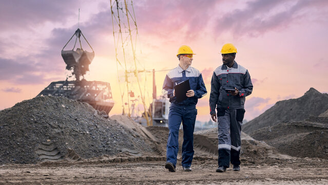 Caucasian male and african american man wearing protective uniform using tablet on mining port. Inspecting open mine, excavator loading sand into truck