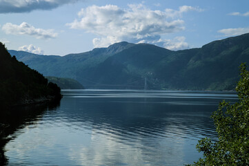 Fjord surrounded by lush green mountains, with a suspension bridge in the distance.