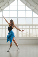 A woman performs a contemporary dance in a bright studio with large windows. Her pose highlights...
