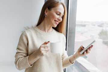 Pretty redhead millennial female in casual clothes standing next to window drinking coffee and...