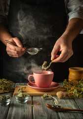 Hands carefully add beneficial herbs to a steamy pink cup surrounded by dried plants, creating a calm kitchen atmosphere inviting a moment of relaxation and tranquility