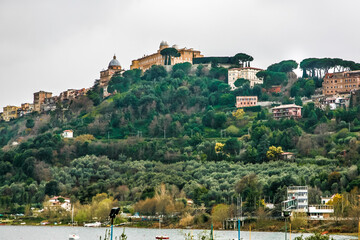 Obraz premium A hill with a large building on top and a small town below papal residence in Italy. The hill is covered in trees and the town is surrounded by water