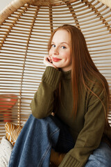 Portrait of pensive ginger-headed cute girl with long hair in green warm jumper and jeans sitting with hands on knees, touching her chin looking up, making wish or dream about falling in love