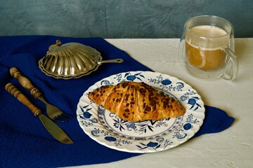 A breakfast scene featuring a croissant with chocolate chips on a decorative blue and white plate, paired with a cup of coffee in a transparent double-walled mug.