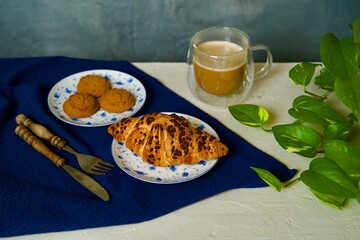 A breakfast scene featuring a croissant with chocolate chips on a decorative blue and white plate, paired with a cup of coffee in a transparent double-walled mug.