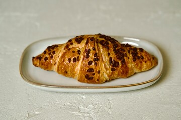 A breakfast scene featuring a croissant with chocolate chips on a simple grey plate on a light textured surface.