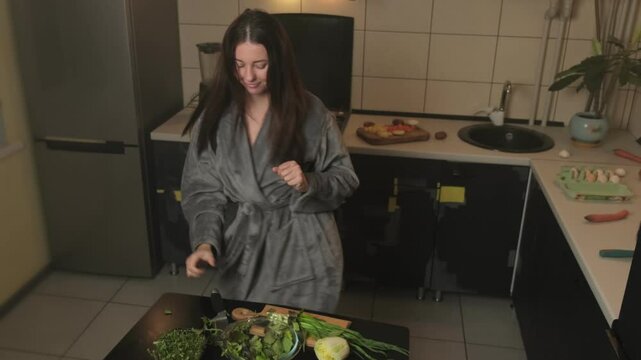 Active young woman in a robe with good mood prepares vegetarian dinner in the kitchen, tossing salad greens into a bowl and dancing joyfully with knife