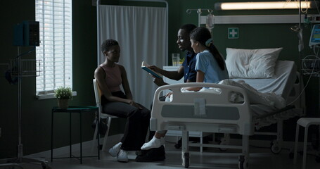 African father and child sit on a hospital bed reading a book together. A mother sits with her child, and the family spends time together while the child is recovering from a serious illness.