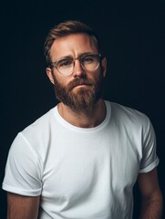 Obraz premium A stylish man with a beard, wearing a plain white t-shirt and trendy clear glasses, posed naturally against a stark black background.