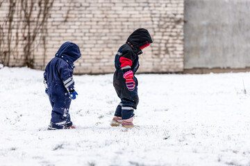 Children playing in winter snow. Two children dressed in warm snowsuits explore the snowy ground near a brick wall, enjoying the crisp winter day in cozy outdoor gear.
