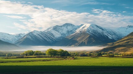 Fototapeta premium Snow-capped mountains with a crisp blue sky, lush green valley beneath, early morning mist rising