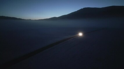 Car with headlights riding through snow covered fields road during sunrise aerial view. Automobile traveling along rural road in winter tracking drone shot - Powered by Adobe