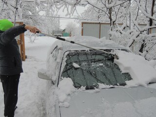a man to the left of the car cleans the windshield from snow with a scraper on a long handle, cleaning a personal car from a layer of snow after a snowfall against the background of an open gate