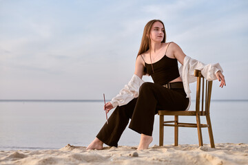 Young female painter sits on wooden chair on sandy beach, holding paintbrush. Serene ocean...