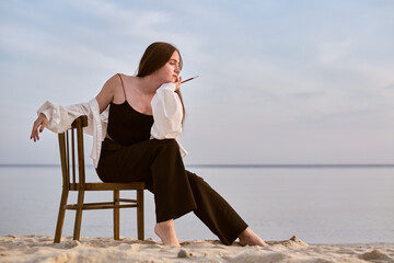 Young female painter sits thoughtfully on beach, gazing at serene sea. Setting sun casts warm glow,...