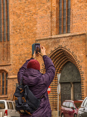 A female tourist wearing a colorful jacket photographs a historic brick building in an urban area, with her smartphone.