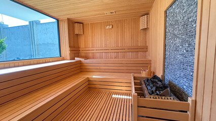 Modern wooden sauna interior featuring polished wood benches, a bucket with a ladle, and a stone heater, illuminated by natural light, perfect for relaxation, wellness, and spa concepts.